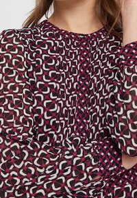 Blusa transparente con un patrón geométrico en tonos de rosa, negro y blanco. Detalles de plisado y botones decorativos en el escote.