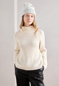 Pull col montant côtelé crème avec des manches longues, associé à un pantalon noir. Le pull présente une coupe décontractée et une texture douce.