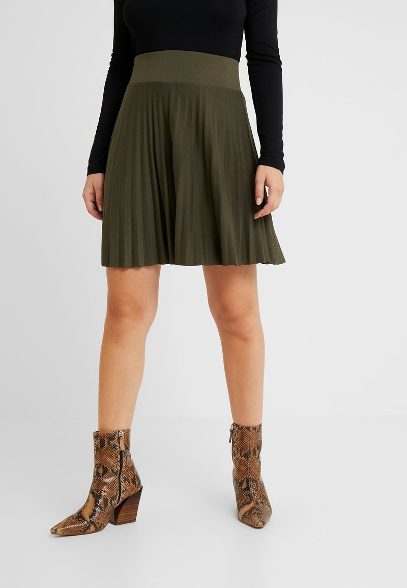 Jupe plissée vert olive avec une large ceinture, associée à des bottines à talons hauts en imprimé serpent marron avec un bout pointu.