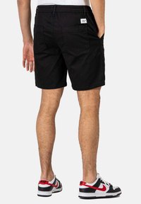 Pantalones cortos negros con una cinturilla elástica, dos bolsillos traseros y una pequeña etiqueta de la marca. Se llevan con zapatillas negras y blancas con detalles en rojo.