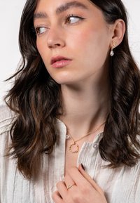 Gold-colored hoop necklace with a pendant, delicate earrings with a pearl drop, and a gold ring adorned with small stones, worn on a textured blouse.