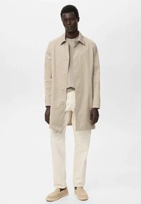 Beiger Baumwoll-Trenchcoat mit Knopfleiste, getragen über einem hellgrauen Hemd, kombiniert mit cremefarbenen Hosen und beigen Slippern.