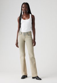 Jean beige clair avec un design évasé, associé à un débardeur blanc ajusté. Le modèle porte des bottes à talons noires ; coiffure minimale, couleurs neutres.