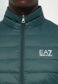 Muškarac nosi teal jaknu s kvadratićima zatvorenu do kraja, s bijelim EA7 Emporio Armani logotipom na lijevoj strani prsa.