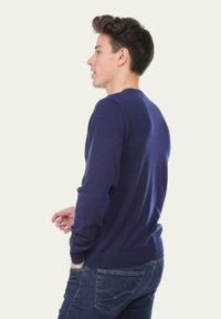 Un jeune homme aux cheveux bruns porte un pull bleu marine et un jean bleu, se tenant de profil devant un fond clair.
