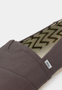 Καφέ καμβάς παπούτσι slip-on με εσωτερική σόλα με ζικ ζακ σχέδιο και ορατή πλαϊνή ετικέτα με την ένδειξη "TOMS."