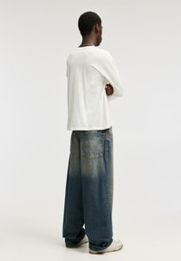 Camiseta blanca de manga larga combinada con vaqueros azules desteñidos y oversize. Los vaqueros presentan un efecto degradado y dos bolsillos traseros. Se ven zapatos blancos.