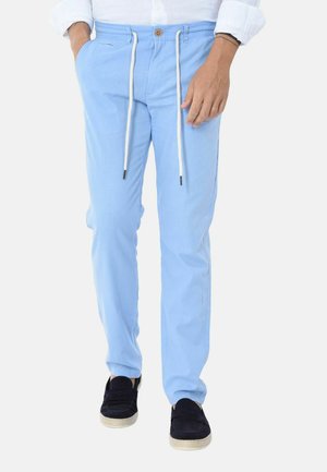 Uomo con pantaloni azzurri chiaro con coulisse, camicia bianca con maniche rimboccate e scarpe nere slip-on, in piedi con le mani in tasca.