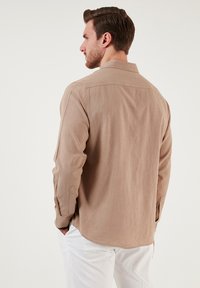 Camicia beige a maniche lunghe in un tessuto testurizzato con colletto classico. Presenta una vestibilità rilassata e polsini con bottone, abbinata a pantaloni bianchi.