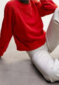 Personne portant un sweat rouge avec une broderie florale sur les épaules et un pantalon blanc, assise sur un sol gris.