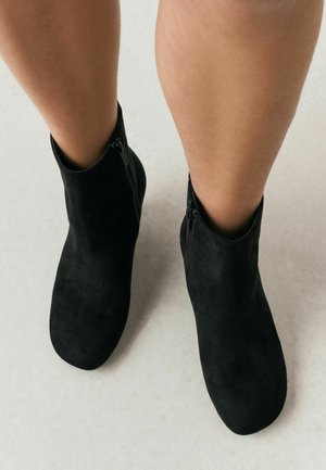 Legs wearing black suede ankle boots with side zippers standing on a light floor.