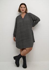 Robe imprimée en noir et blanc avec des manches longues, décolleté en V, poches latérales, coupe ample, portée avec des bottines noires. Texture de tissu lisse.