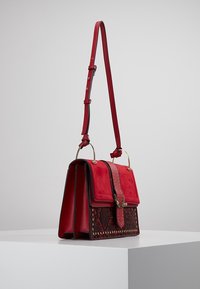 Bolso cruzado de cuero rojo y ante con correa ajustable, asas en forma de anillo dorado, solapa con patrón de serpiente tachonado y cierre decorativo en forma de pájaro dorado.