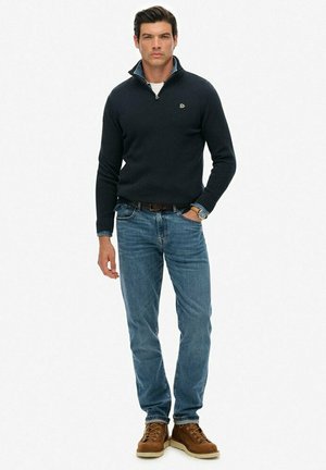 Hombre de pie vistiendo un suéter oscuro de media cremallera, jeans azules, botas marrones con cordones, reloj en la muñeca izquierda, sobre un fondo blanco.