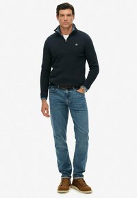 Marineblauer Pullover mit Reißverschlusskragen, getragen über einem weißen Hemd. Blaue Jeans und braune Schnürstiefel vervollständigen das Outfit.