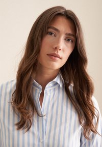 Ung kvinde med langt brunt hår iført blå og hvid stribet skjorte med knapper, neutral ansigtsudtryk, beige baggrund.