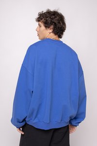 Liela izmēra zila kokvilnas kapuce, ar apaļu kakla izgriezumu, nolaižamām plecu līnijām un ribota apakšmalām, valkāta ar melnām biksēm.