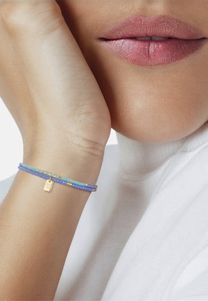 Primer plano de los labios y la muñeca de una persona que lleva dos pulseras de cuentas azules y turquesas con un pequeño charm dorado, sobre un fondo de jersey blanco de cuello alto.