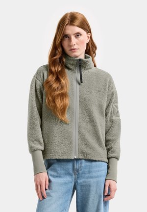 Young woman with long red hair wears gray fleece zip-up jacket and light blue jeans standing against plain background.