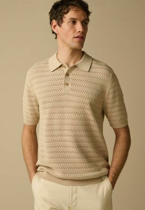 Man met krullend bruin haar, gekleed in een beige korte mouw gebreide poloshirt en lichte broek, handen in de zakken, kijkt naar rechts.