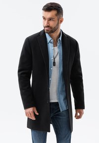 Schwarzer Wollmix-Mantel mit Reverskragen, zwei Fronttaschen und einer Knopfverschluss, über ein Jeanshemd und ein weißes T-Shirt geschichtet.
