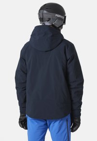 Námořnická vodotěsná lyžařská bunda s kapucí, s hladkou texturou a nastavitelnými manžetami, nošena přes modré lyžařské kalhoty a černé rukavice.