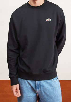 Mann trägt schwarzen Sweatshirt mit Rundhalsausschnitt und kleinem rot-weißem Sneaker-Patch auf der linken Brust, kombiniert mit hellblauen Jeans.