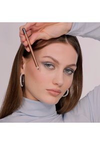 En makeuppenna i handen, en modell med långt brunt hår bär en ljusblå polotröja och stora cirkulära silverörhängen, och visar upp en sotig ögonmakeup.