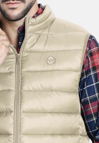 Chaleco acolchado beige con cuello alto, cierre de cremallera y un logotipo circular. Lleva encima una camisa de franela a cuadros multicolor con texturas visibles.