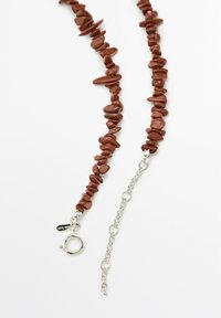 Bruine stenen kralenketting met onregelmatige vormen en een zilveren ketting. Beschikt over een ronde sluiting met verstelbare schakels voor de lengte.