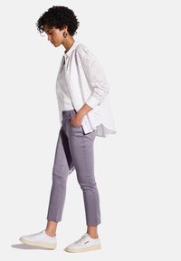 Camicia bianca in pizzo, pantaloni grigi, sneakers bianche. La camicia presenta un intricato design a taglio, mentre i pantaloni hanno una vestibilità sartoriale e sono alla caviglia.