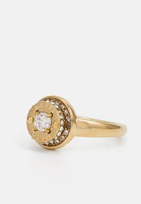 Zlatni prsten s okruglim središnjim dijelom koji sadrži prozirni kamen, oko kojeg su raspoređeni mali rhinestones. Ugravirani naziv branda "GUESS" na središnjem dijelu.