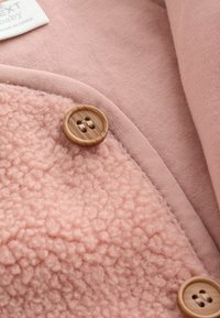 Világos rózsaszín baba ruházat, amely puha fleece-textúrával, sima anyaggal és díszítő, természetes fa megjelenésű kerek gombbal rendelkezik.