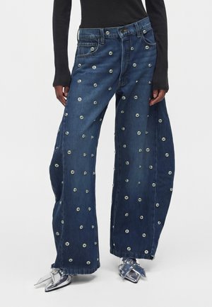 Jeans blu a gamba larga con decorazioni in occhielli argentati indossati con scarpe metalliche argentate con punta e una maglia nera a maniche lunghe.