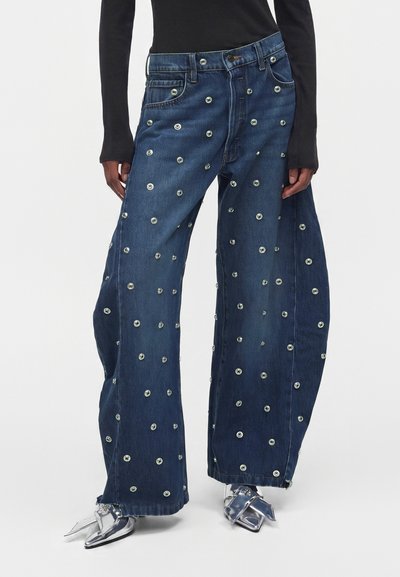 Jean bleu à jambes larges avec des œillets argentés, porté avec des chaussures pointues métalliques argentées et un haut noir à manches longues.