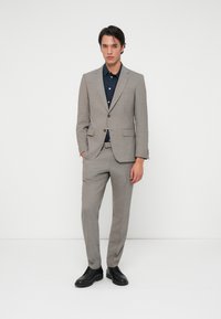 Ανοιχτό γκρι κοστούμι από υφή υφάσματος με κομψή εφαρμογή, συνδυασμένο με ναυτικό πουκάμισο και μαύρα κοστούμια παπούτσια. Στηρίζεται σε λευκό φόντο.