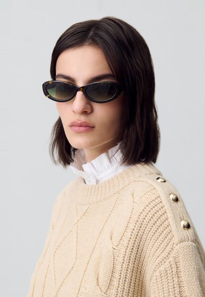 Occhiali da sole con una forma ovale elegante, lenti scure e rifiniture in tartaruga. Indossati con un maglione in crema a trecce con bottoni dorati.