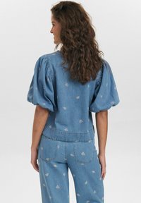 Ljusblå denimtop med puffärmar, prydd med blommönster. Avslappnad passform, sömmar som detaljer, och en fåll som slutar vid midjan.