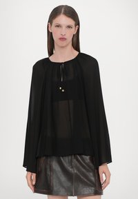 Black sheer blouse with long cape-like sleeves, gathered neckline, and a tie closure. Features gold accents and a flowing, lightweight fabric.