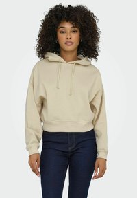 Beige verkürzter Kapuzenpullover mit Kordeln, übergroßer Passform, gerippten Bündchen und glatter Textur, kombiniert mit dunklen Jeans.