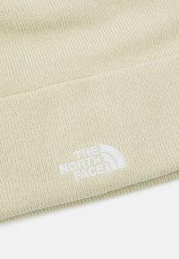 Strikket hue i en lys beige farve med et struktureret mønster og et hvidt broderet logo, "The North Face," på forsiden.