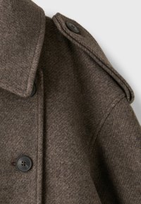 Gros plan sur l'épaule d'un manteau en laine marron avec une épaulette boutonnée et de grands boutons noirs sur la patte de boutonnière avant.