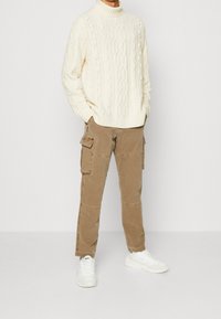 Witte kabelgebreide turtleneck trui, tan kleurige cargo broek met zakken, gecombineerd met witte sneakers. Focus op de brei textuur en het casual ontwerp.