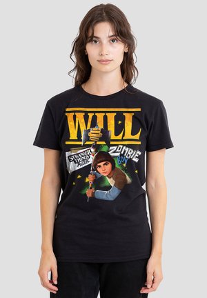 Young woman wearing a black "Will" Stranger Things t-shirt featuring character holding axe with text "Zombie Boy" on a plain background.