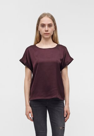 Jeune femme aux cheveux blonds lisses portant un chemisier en satin violet foncé à manches courtes et un jean noir déchiré, debout devant un fond uni.