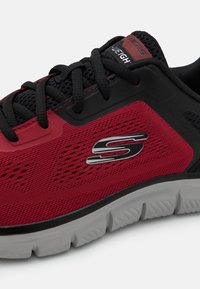 Röd och svart atletiksko med ett ventilerande meshöverdrag, texturerade accenter, vadderad krage och en flexibel grå sula. Har logotypdetalj.