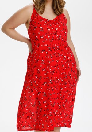 Femme portant une robe rouge sans manches avec de petits motifs floraux blancs et noirs, main posée sur la hanche, fond blanc.