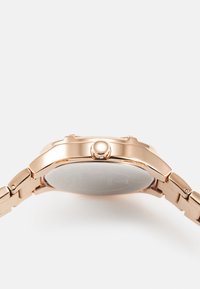 Montre en or rose avec un bracelet métallique, boîtier rond, couronne sur le côté droit, et un fond en verre transparent montrant le mécanisme intérieur.