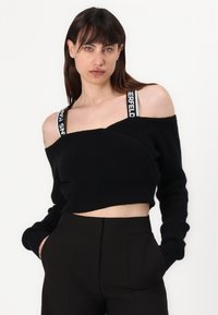 Schwarzer, gestrickter Crop-Pullover mit schulterfreiem Design, geripptem Muster und kontrastierenden Trägerdetails. Kombiniert mit schwarzen, hochtaillierten Hosen.