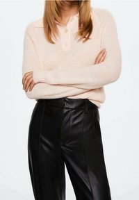 Femme portant un pull en maille rose pâle avec des boutons, les bras croisés, associé à un pantalon en cuir noir taille haute devant un fond blanc.